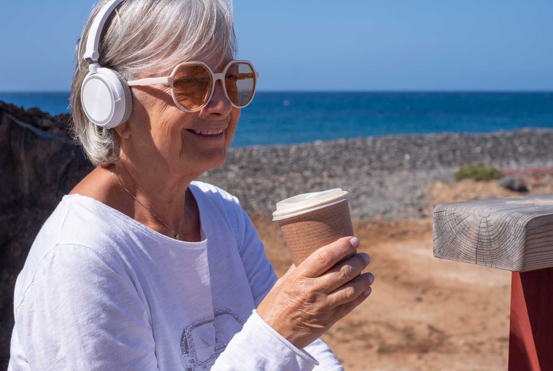 Senior Woman Enjoying Coffee and Listening to Music with Headphones at the Beach