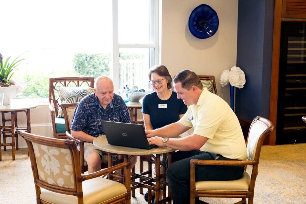 Two seniors sit with their caregiver as he shows them life plan communities on the computer 